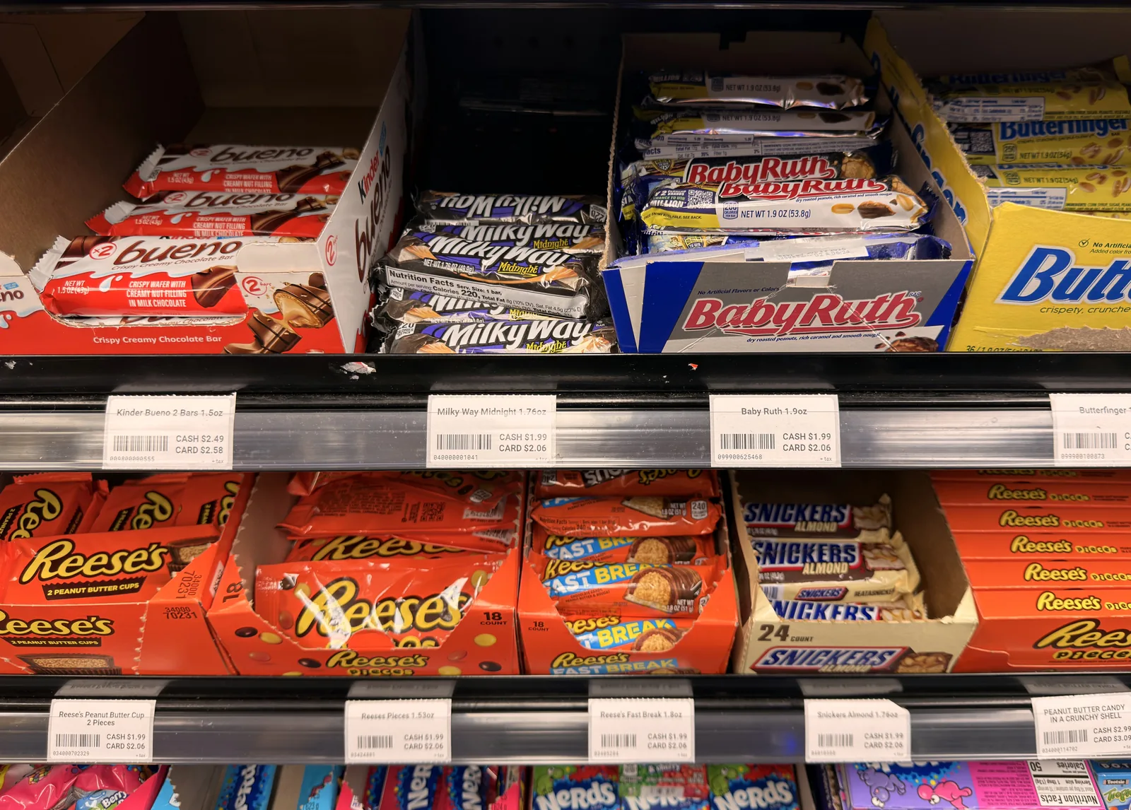 Printed shelf labels beneath candy products, each showing the product name, barcode, cash price and card price